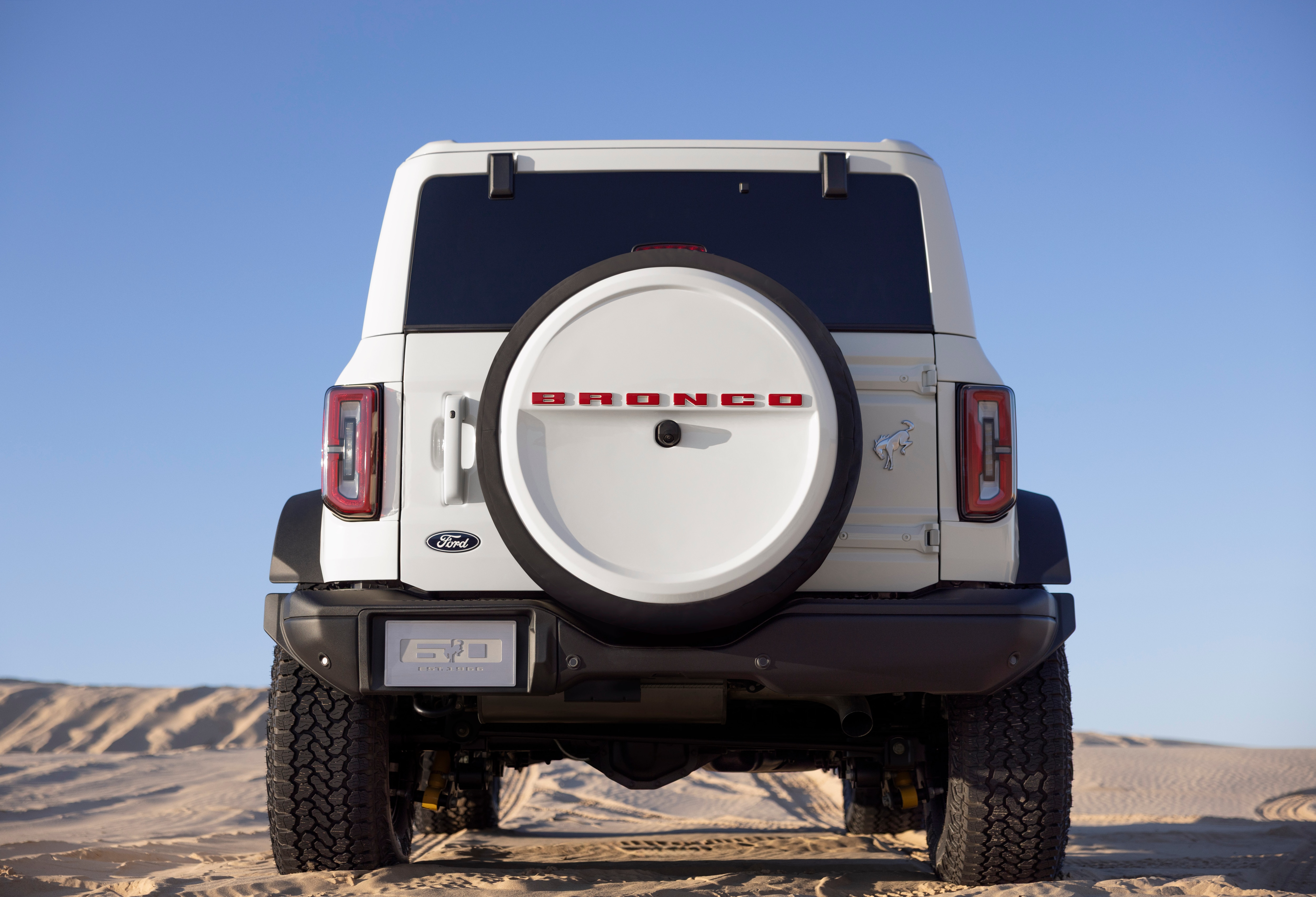 2026 Ford Bronco 60th Anniversary Package front view showing a white vehicle with "BRONCO" across the rear in red letters. Vehicle is on the sand with a blue sky overhead.